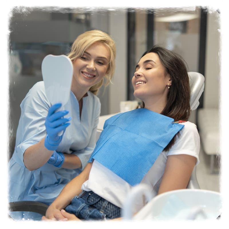 Na cadeira de dentista, paciente vê o resultado do seu tratamento, por um espelho segurado pela dentista que a tratou, ambas com expressão de alegria.