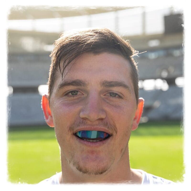 Rapaz no gramado de um estádio de futebol, num dia ensolarado, sorrindo para a câmera, mas o que chama atenção é o seu protetor bucal colorido.