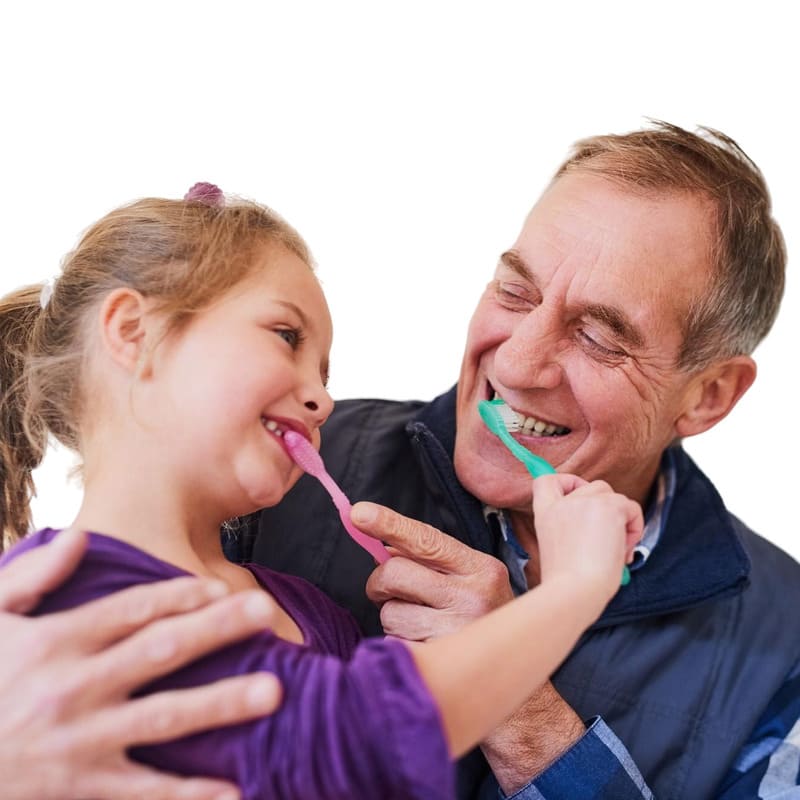 Um idoso e uma criança escovando os dentes um do outro, numa brincadeira com risadas e um segurando a escova de dentes na boca do outro.