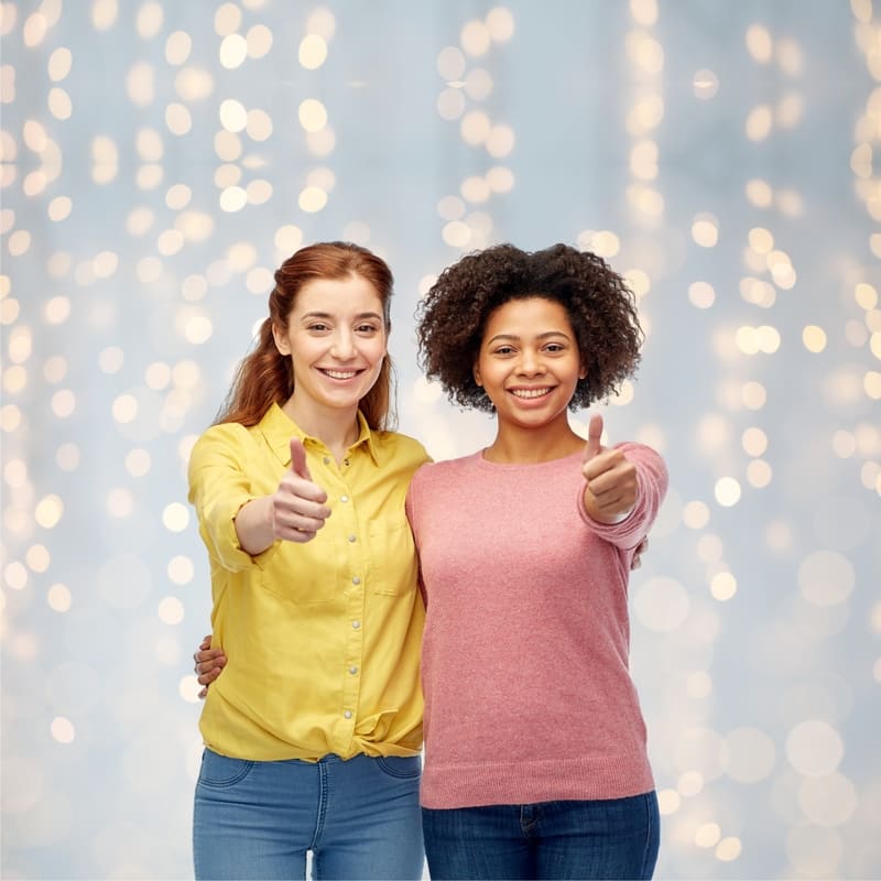 Duas moças, amigas, uma morena e outra ruiva, num ambiente iluminado, e ambas sorrindo, abraçadas lado a lado, e com o outro braço estendido e com um sinal de positivo com o polegar.