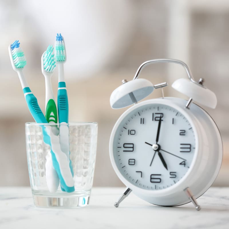 Numa pia de banheiro, um copo com escovas de dentes, ao lado de um relógio despertador, simbolizando a pressa com os horários não deve impedir os cuidados odontológicos.