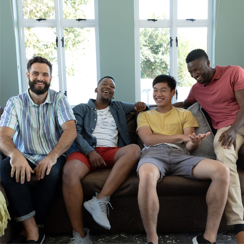 Grupo de quatro amigos conversando e sorrindo, sentados em um sofá, numa sala iluminada e com uma janela atrás, que mostra um local arborizado.