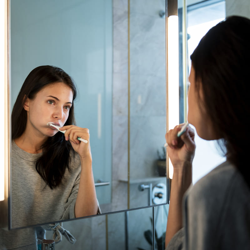 Mulher morena no banheiro enfrente ao espelho, escovando os dentes, com expressão de cansada e de quem não queria estar fazendo isso.