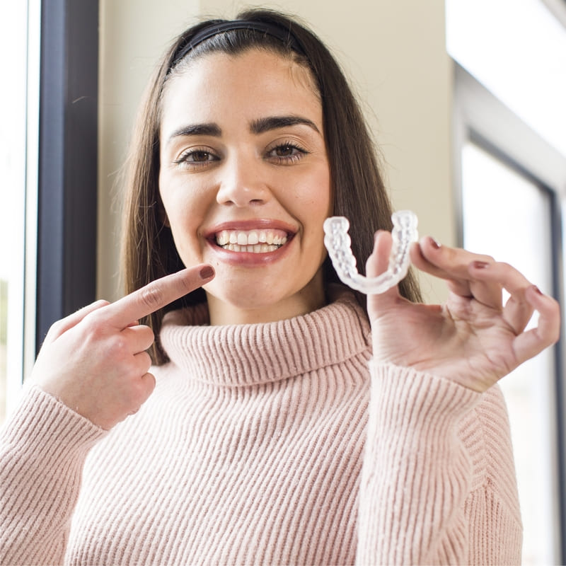 Uma moça de cabelos castanhos segura o aparelho Invisalign, e aponta para o seu sorriso, mostrando o bom resultado.
