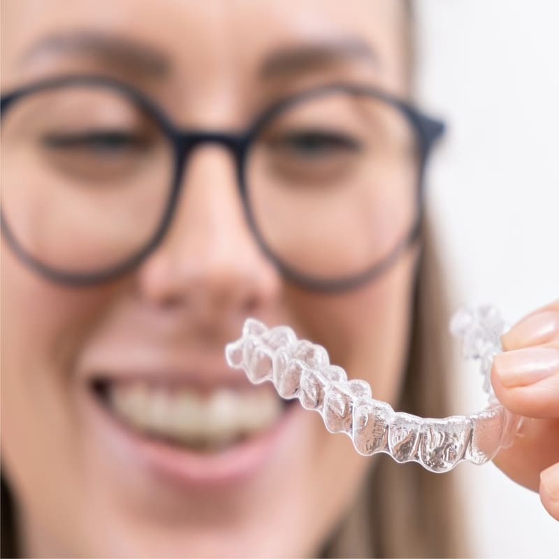 Uma moça, de óculos, num segundo plano mais ao fundo e desfocado, segura o aparelho dental mais próximo a câmera fotográfica, permitindo um bom foco no aparelho e nos dedos que o seguram.