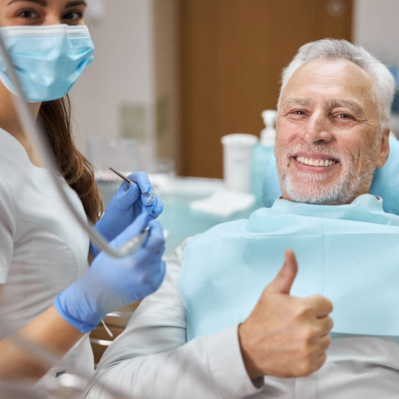 Um paciente idoso sentado na cadeira de dentista, sorrindo e fazendo sinal de positivo, assim como a dentista ao lado dele, ambos olhando para a câmera.