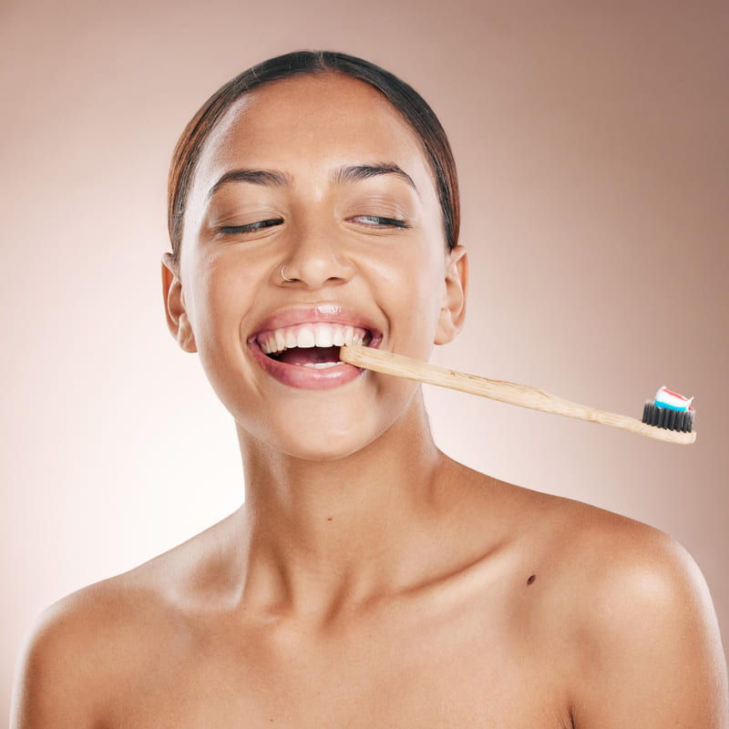 Uma moça sorridente, mordendo a ponta do cabo de uma escova de dentes, e olhando para a outra extremidade da escova, onde há pasta de dente colocada.