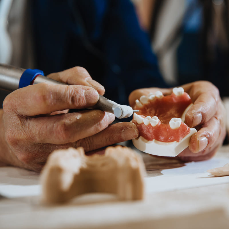 Um protético, em sua bancada de trabalho, ajusta uma prótese com um motor de dentista.