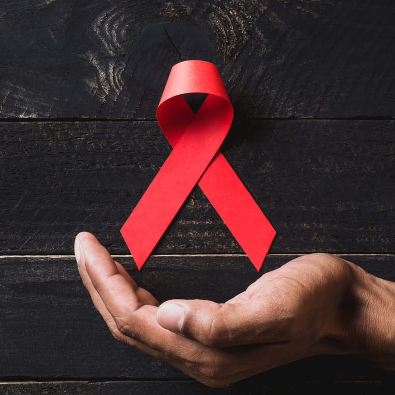Com um fundo de madeira na cor preta, uma mão abaixo, em forma de concha, simula um suporte para o laço vermelho característico da luta contra o HIV / AIDS