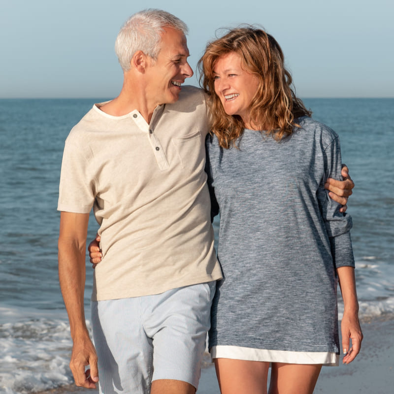 Casal de terceira idade, representando aposentados, sorrindo e caminhando numa praia ensolarada.