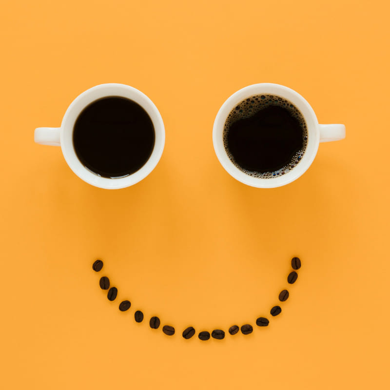 Em uma mesa de cor laranja, duas xícaras de café lado a lado, como que formando dois olhos, e abaixo uma fileira de grãos de café, de forma curva, formando uma boca sorrindo.