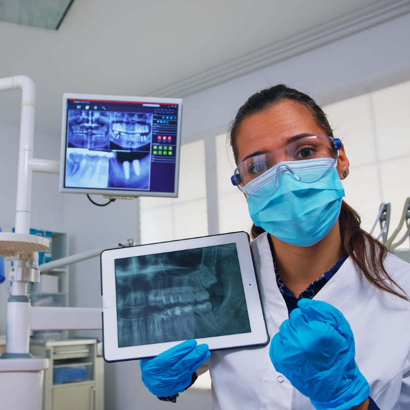 Uma dentista em seu consultório, com duas telas abertas, uma no monitor mais ao fundo e outra de um tablet, na mão da dentista, mostrando o quanto a tecnologia avançou e trouxe precisão aos diagnósticos odontológicos.