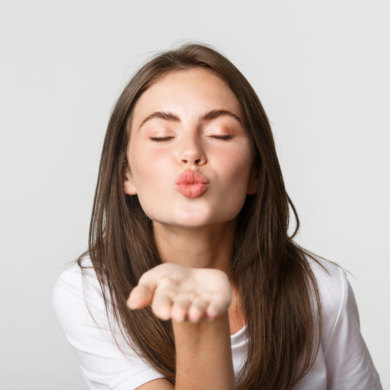 Uma moça jovem, de olhos fechados, expressão de riso suave, fazendo biquinho com a boca, como um beijinho, e a palma da mão extendida à frente como que jogando o beijo para alguém.