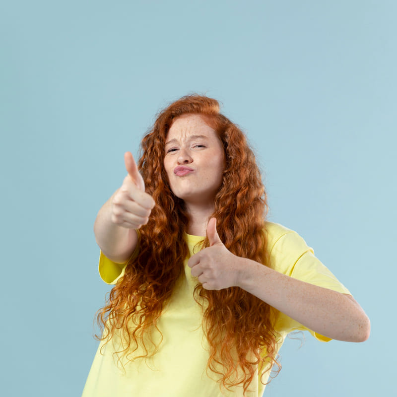 Uma moça ruiva, aparentando 20 anos, com cabelos compridos e ondulados, com camiseta amarela e um fundo azul claro, fazendo sinal de positivo com os dedos e fazendo uma mistira de bico com sorriso, num momento de descontração.